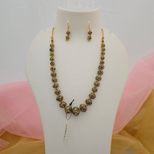 Black and gold beaded necklace mala featuring graduated spherical beads with intricate antique gold-tone patterns and matching black and gold earrings displayed on a white jewellery bust with pink and mustard fabric